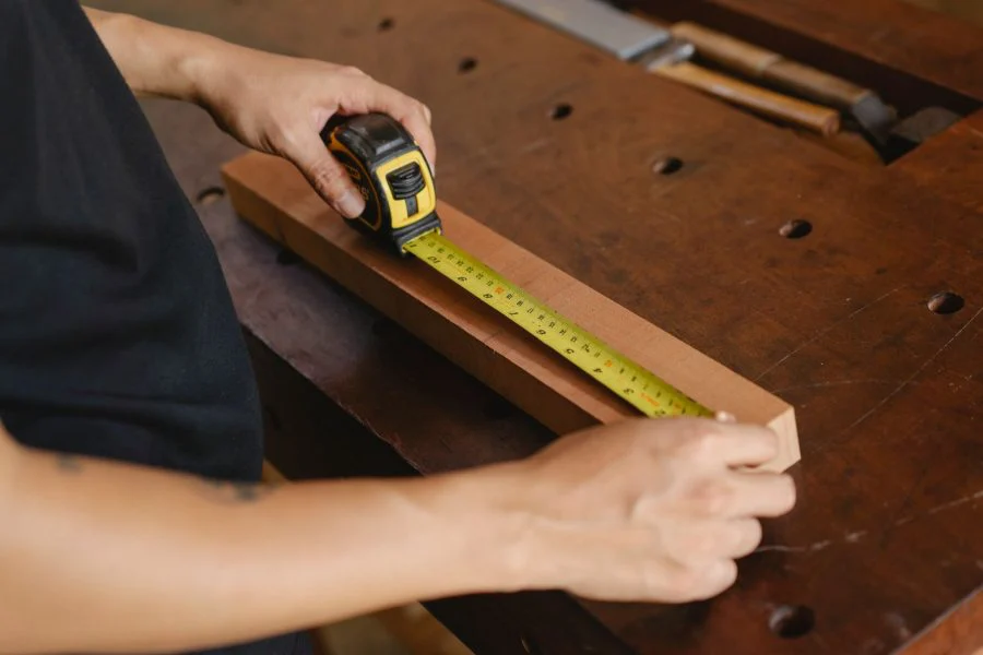 Precise unrecognizable male carpenter accurately measuring wooden detail while using ruler during work at workbench with instruments in professional studio